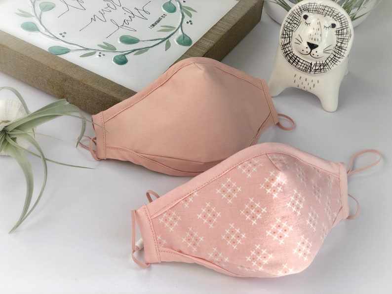 Op de afbeelding: Twee roze gezichtsmaskers met verstelbare oorlussen. E&eacute;n masker is effen roze en het andere heeft een wit en roze kruispatroon. De maskers zijn gemaakt van stof en zijn ontworpen om de neus en mond te bedekken.