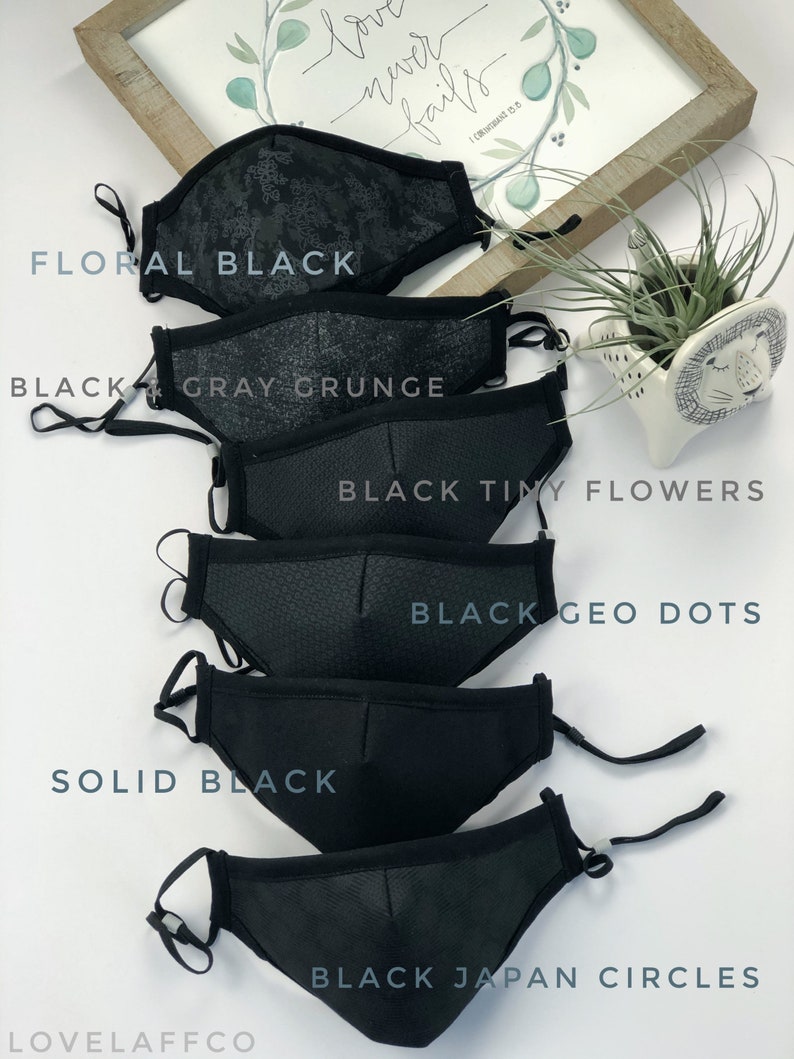 May include: Six black face masks with different patterns. The masks are arranged in a stack, with the top mask being a floral pattern, followed by a gray grunge pattern, a tiny flower pattern, a geometric dot pattern, a solid black mask, and a black mask with small circles. The text "Love never fails" is visible in the background.