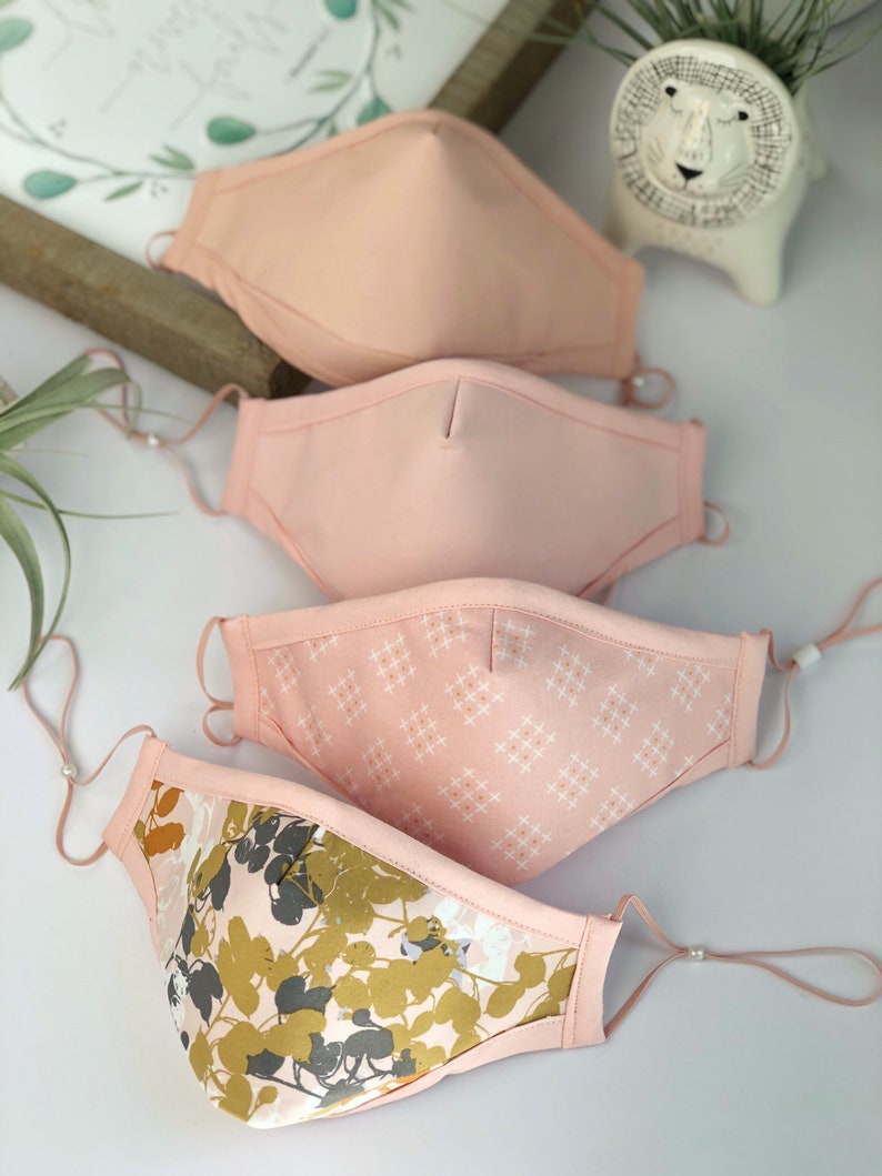 Op de afbeelding: Vier gezichtsmaskers in roze en perziktinten, met verstelbare oorlussen. E&eacute;n masker heeft een wit rasterpatroon en een ander heeft een bloemenontwerp met gouden en grijze bladeren. De maskers zijn gemaakt van stof.
