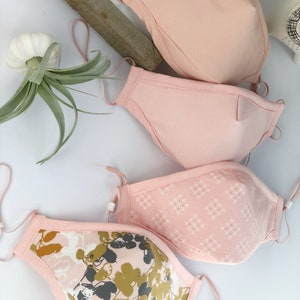 Op de afbeelding: Vier gezichtsmaskers in roze tinten, met verstelbare oorlussen. E&eacute;n masker heeft een bloemenpatroon in olijfgroen, grijs en wit. Een ander heeft een klein wit kruispatroon. De andere twee zijn effen roze.