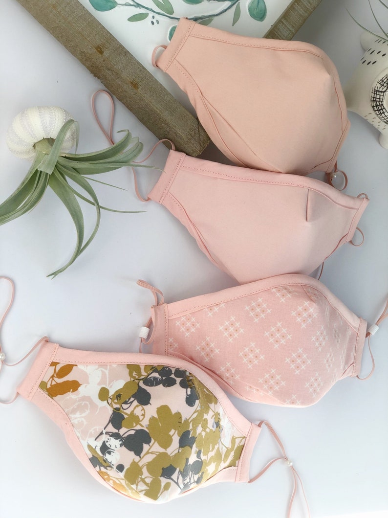 Op de afbeelding: Vier gezichtsmaskers in roze tinten, met verstelbare oorlussen. E&eacute;n masker heeft een bloemenpatroon in olijfgroen, grijs en wit. Een ander heeft een klein wit kruispatroon. De andere twee zijn effen roze.