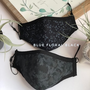May include: Two black face masks with floral patterns. The top mask has a blue floral pattern and the bottom mask has a gray floral pattern. The masks are labeled "Blue Floral Black" and "Black & Gray Floral".