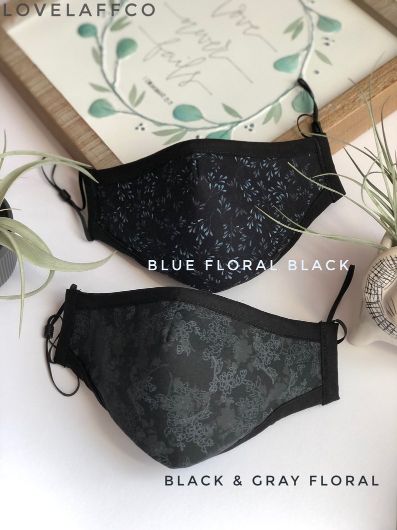 May include: Two black face masks with floral patterns. The top mask has a blue floral pattern and the bottom mask has a gray floral pattern. The masks are labeled "Blue Floral Black" and "Black & Gray Floral".