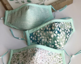 Op de afbeelding: Drie stoffen mondmaskers met verschillende patronen. De maskers zijn allemaal lichtblauw met een witte achtergrond. Het eerste masker heeft een geometrisch patroon, het tweede heeft een bladpatroon en het derde heeft een bloemenpatroon.