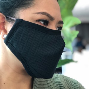 May include: A black face mask with a textured pattern. The mask is worn by a person with dark hair and is secured with elastic ear loops.