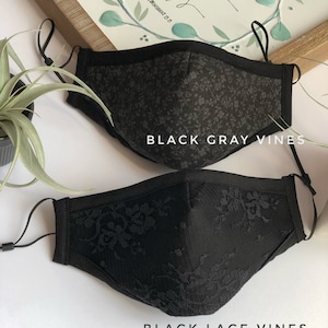 May include: Two black face masks with floral patterns. The top mask has a gray floral pattern and the bottom mask has a black lace pattern. Both masks have black elastic ear loops.