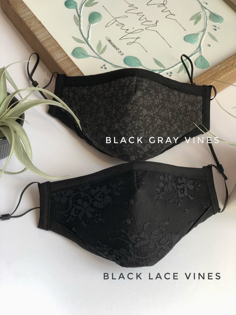 May include: Two black face masks with floral patterns. The top mask has a gray floral pattern and the bottom mask has a black lace pattern. Both masks have black elastic ear loops.