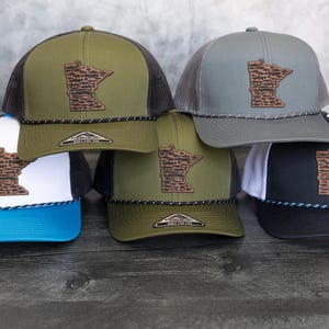 May include: Five different trucker hats with a Minnesota state outline patch. The hats are in various colors, including green, gray, white, and black. The patches are all brown and feature a detailed outline of the state.