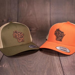 May include: Two trucker hats with a brown leather Wisconsin state outline patch with a fish scale pattern. One hat is green with a tan mesh back and the other is orange with a tan mesh back.
