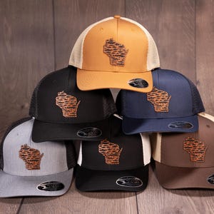 May include: Six different color baseball caps with a brown leather patch of the state of Wisconsin on the front of each cap. The caps are arranged in a pyramid shape.