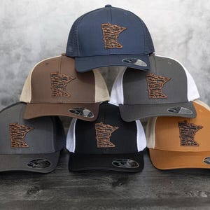 May include: Six different baseball caps with a brown leather Minnesota state outline patch on the front. The caps are in various colors, including black, brown, gray, tan, and blue.