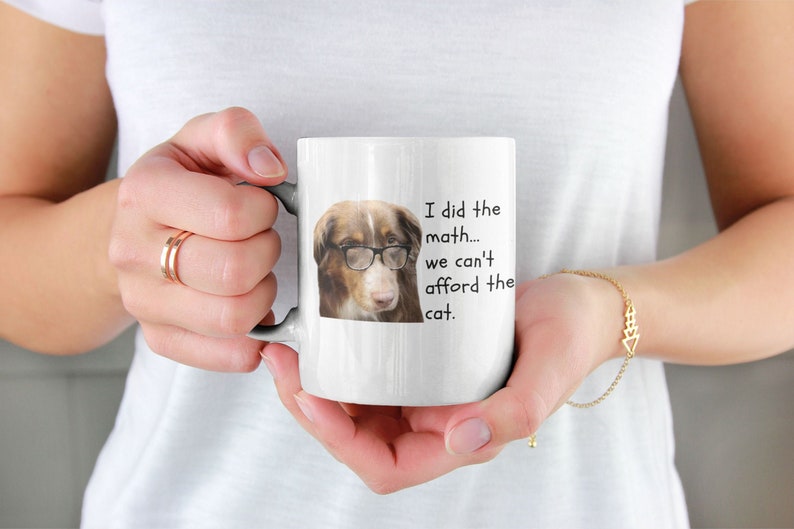 Puede incluir: Taza de cer&aacute;mica blanca con un perro marr&oacute;n y blanco que lleva gafas. La taza tiene el texto "Hice las cuentas... no podemos permitirnos el gato."