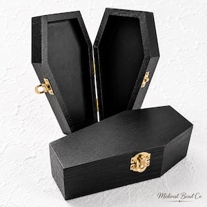 May include: Two black wooden boxes shaped like coffins. One box is standing open, revealing a hollow interior, while the other is closed in the foreground. Both feature gold-colored metal latches and hinges. The text Midwest Bead Co is visible.