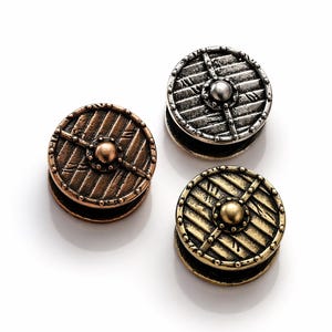 May include: Three small, round, metal shield-shaped objects in bronze, silver, and gold tones. Each shield has a central boss and a cross design, with a textured, weathered appearance. The shields are likely decorative or functional.