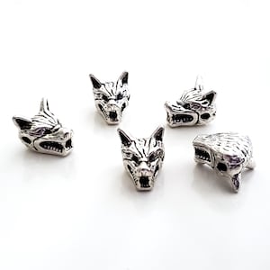 May include: Five silver-colored wolf head beads with detailed facial features, including open mouths and sharp teeth. The beads are scattered on a white surface, showcasing their intricate design and metallic sheen.