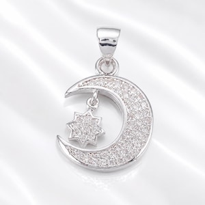 May include: A silver-toned metal pendant shaped like a crescent moon with a small eight-pointed star hanging from the center. The entire surface of the moon and the star is covered in small, clear, sparkling stones, and it has a bail at the top.