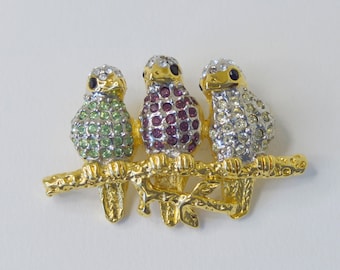 May include: Gold tone brooch featuring three jeweled birds perched on a branch. The birds are adorned with green, purple, and clear rhinestones.
