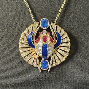 May include: Gold pendant necklace with a scarab beetle design. The pendant features blue and red details, clear crystals, and a gold chain. The necklace is set against a dark background.