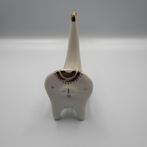 May include: A white ceramic ring holder shaped like a tooth. It features a tall, gold-tipped cone for the ring and a face design with gold and brown details. The base is tooth-shaped. The holder is likely for a dressing table.