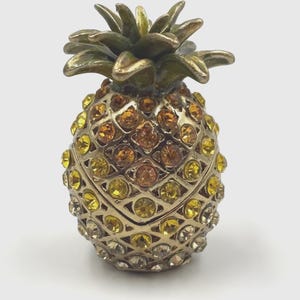May include: A decorative pineapple-shaped trinket box featuring a metallic gold-tone finish and a textured surface encrusted with numerous faceted amber, yellow, and clear crystals. The top of the pineapple has a sculpted green metal leaf crown.