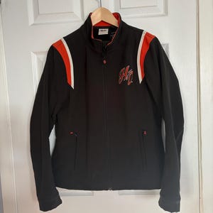 May include: Black zip-up jacket with orange and white shoulder accents. Features a high collar and a red logo on the chest. The jacket has two front pockets and a full-length zipper.