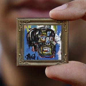 Może przedstawiać: Miniaturowe, oprawione dzieło sztuki z kolorowym, abstrakcyjnym wzorem czaszki. Obraz ma niebieskie tło z czarnymi, żółtymi i czerwonymi akcentami. Rama jest złota i ozdobna, dodając dziełu charakteru vintage.