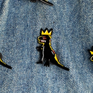 May include: Black dinosaur enamel pin with a yellow crown and yellow and red accents. The pin is set against a blue denim background. The dinosaur is wearing a gold crown.