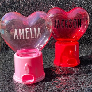 May include: Two heart-shaped gumball machines. One is pink with the name "AMELIA" in white, and the other is red with "JACKSON" in black. Both have a clear, bubble-like top and a base with a dispensing slot.