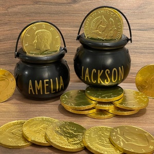 May include: Two black cauldron-shaped containers with gold lettering, one reading "AMELIA" and the other "JACKSON", are filled with gold coins. Additional gold coins are scattered around the cauldrons on a wooden surface. The coins are a bright gold color.