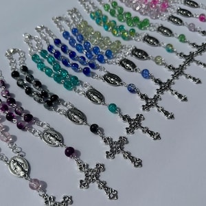 May include: A collection of rosaries with colorful beads in shades of pink, purple, black, green, blue, and clear. Each rosary features a silver-toned crucifix and a centerpiece with a religious image. The beads are strung on a silver chain.