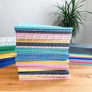 Può includere: Una pila di quaderni colorati con diversi motivi geometrici. I quaderni sono disposti a forma di arcobaleno, con quaderni rosa, gialli, blu, verdi e neri.