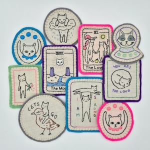 May include: A collection of ten embroidered fabric patches featuring various cat illustrations. Designs include tarot-style cards with text like The Moon and The Lovers, a cat in a UFO, a cat riding a bird with text LETS GO, and a cat lifting another cat with text I AM STRONG.