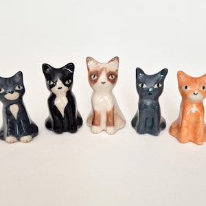 May include: A set of five small ceramic cat figurines standing in a row. The cats feature various coat patterns including a grey tabby, a black and white tuxedo, a tan and white point, a solid dark grey, and an orange tabby with distinct painted faces.