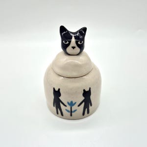 May include: A small, off-white ceramic jar with a lid topped by a black and white cat head figurine. The jar body features a hand-painted design of two black cat silhouettes standing on either side of a small blue flower on a speckled surface.