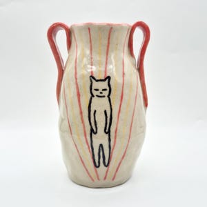 May include: A ceramic vase with two red handles, featuring a speckled off-white glaze. The body is decorated with vertical yellow and red painted stripes radiating from a central black line drawing of a standing cat with a simple facial expression.