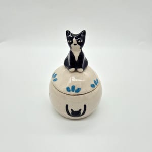 May include: A small, round, cream-coloured ceramic jar with a lid featuring a sculpted black and white cat figurine on top. The jar is decorated with hand-painted blue leaf-like accents and a black cat silhouette graphic on the front surface.
