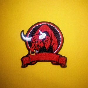 Puede incluir: Parche de toro rojo bordado con un anillo de nariz blanco y una pancarta roja. El toro está dentro de un círculo negro con un contorno blanco.