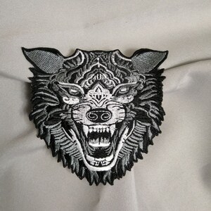 Pode incluir: Patch bordado com um design detalhado de cabeça de lobo. O patch mostra um lobo rosnando com dentes afiados, em preto, cinza e branco. O design tem detalhes intrincados, ideal para adicionar a roupas ou acessórios.