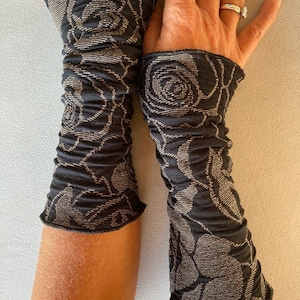 Könnte beinhalten: Ein Paar schwarz-goldene Armstulpen mit floralem Muster. Die Armstulpen sind aus einem weichen, strukturierten Stoff gefertigt und haben ein gerafftes Design. Das florale Muster ist hellgoldfarben.