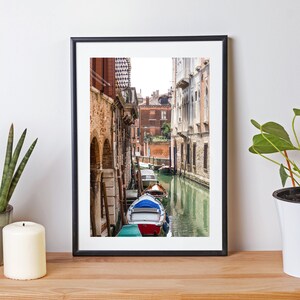 Puede incluir: Fotograf&iacute;a enmarcada de un canal veneciano con barcos coloridos y edificios hist&oacute;ricos. La imagen est&aacute; sobre una superficie de madera, con una planta en maceta y una vela.
