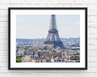 May include: A framed wall art print featuring a high-angle view of the Eiffel Tower rising above the dense cityscape of Paris, France. The iron structure of the tower is dark grey, set against a pale blue sky, overlooking light-coloured buildings.