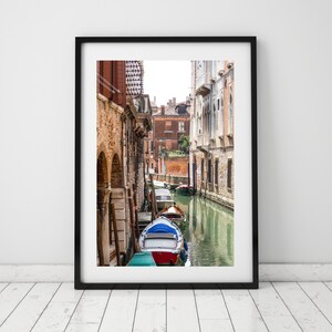 Puede incluir: Una impresi&oacute;n enmarcada de una escena del canal veneciano. La imagen presenta una v&iacute;a fluvial estrecha flanqueada por edificios de ladrillo envejecido. Un peque&ntilde;o bote con detalles rojos, blancos y azules est&aacute; en primer plano, con otros botes m&aacute;s abajo en el canal. El agua refleja los edificios y el cielo.