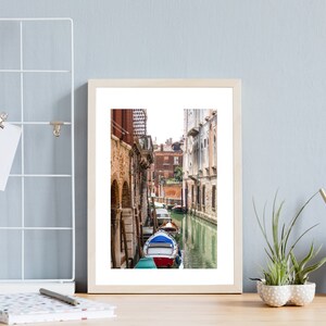 Puede incluir: Una impresi&oacute;n enmarcada de una escena del canal veneciano. La imagen presenta una v&iacute;a fluvial estrecha flanqueada por edificios antiguos con puertas arqueadas. Una g&oacute;ndola con detalles rojos y azules est&aacute; amarrada en el agua. La impresi&oacute;n est&aacute; en un marco de madera clara, exhibida en un escritorio.