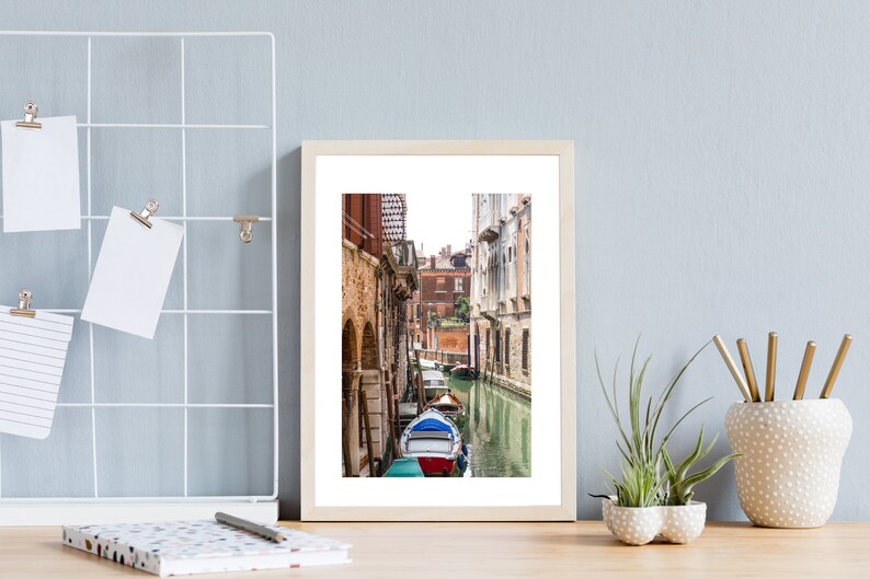 Puede incluir: Una impresi&oacute;n enmarcada de una escena del canal veneciano. La imagen presenta una v&iacute;a fluvial estrecha flanqueada por edificios antiguos con puertas arqueadas. Una g&oacute;ndola con detalles rojos y azules est&aacute; amarrada en el agua. La impresi&oacute;n est&aacute; en un marco de madera clara, exhibida en un escritorio.