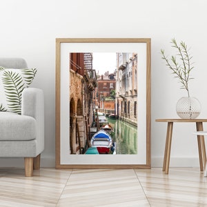 Puede incluir: Una impresi&oacute;n enmarcada de una escena del canal veneciano. La imagen presenta una v&iacute;a fluvial estrecha flanqueada por edificios de ladrillo antiguos. Un barco azul y rojo est&aacute; amarrado en el canal. La impresi&oacute;n se exhibe en una pared blanca.