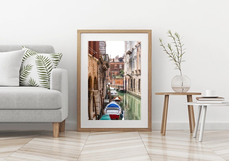 Puede incluir: Una impresi&oacute;n enmarcada de una escena del canal veneciano. La imagen presenta una v&iacute;a fluvial estrecha flanqueada por edificios de ladrillo antiguos. Un barco azul y rojo est&aacute; amarrado en el canal. La impresi&oacute;n se exhibe en una pared blanca.