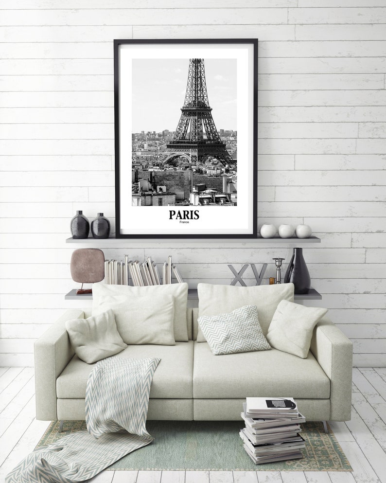 May include: Black and white framed print of the Eiffel Tower in Paris, France. The print is displayed above a light-colored sofa with pillows and a patterned throw. The word "Paris" is printed at the bottom of the print.