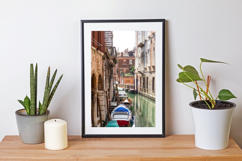 Puede incluir: Fotograf&iacute;a enmarcada de un canal veneciano con barcos coloridos y edificios hist&oacute;ricos. La imagen est&aacute; sobre una superficie de madera, con una planta en maceta y una vela.