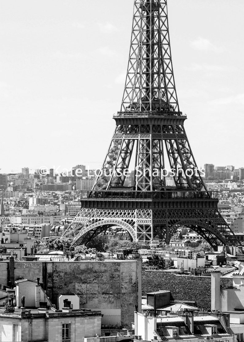 May include: Black and white photograph of the Eiffel Tower in Paris, France. The iconic wrought-iron lattice tower dominates the frame, with the city skyline and buildings in the background. The image is a high-angle shot.