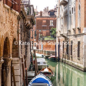 Puede incluir: Una vista pintoresca de un canal veneciano con varios botes amarrados a lo largo del agua. Los edificios a ambos lados son de ladrillo y piedra, con ventanas arqueadas y balcones. El agua es verdosa y el cielo est&aacute; despejado.